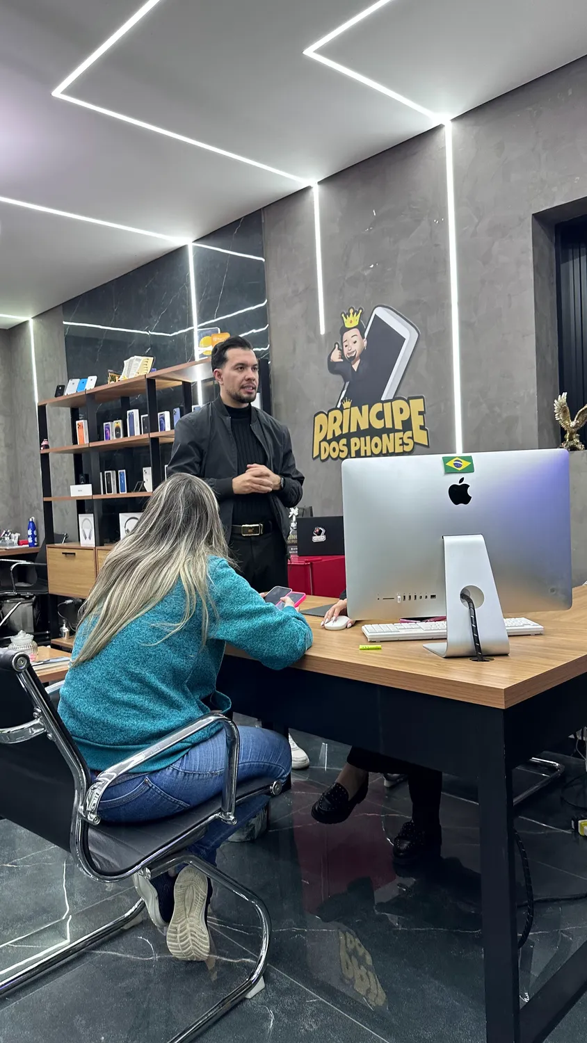 Atendente da Príncipe dos Phones ao lado do painel da marca, atendendo um cliente no balcão com iMac.